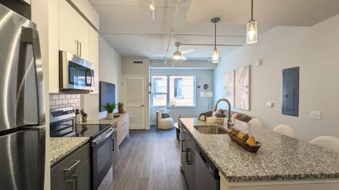 An open-concept layout showing a kitchen with a large granite-style island, stainless steel appliances, and gray lower cabinetry transitioning into the living area.