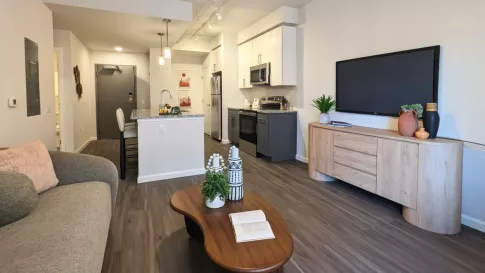 An open-concept layout showing a kitchen with a large granite-style island, stainless steel appliances, and gray lower cabinetry transitioning into the living area.
