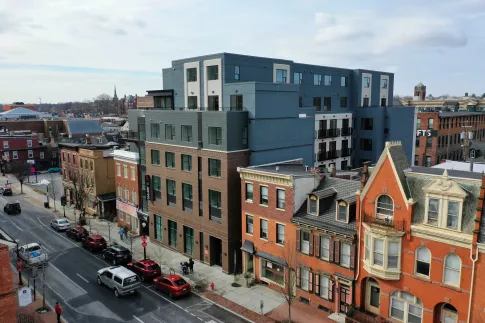 Elevated view of a modern blue multi-story building alongside older brick rowhouses and parked cars on a city street.