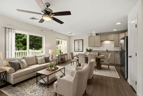 Bright open-plan living room with beige sofa, dining table, and kitchen featuring grey cabinets and stainless steel appliances.