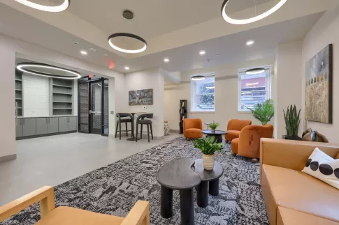 Spacious, modern common lounge with orange armchairs, a brown sofa, high-top seating, and decorative plants under circular lights.