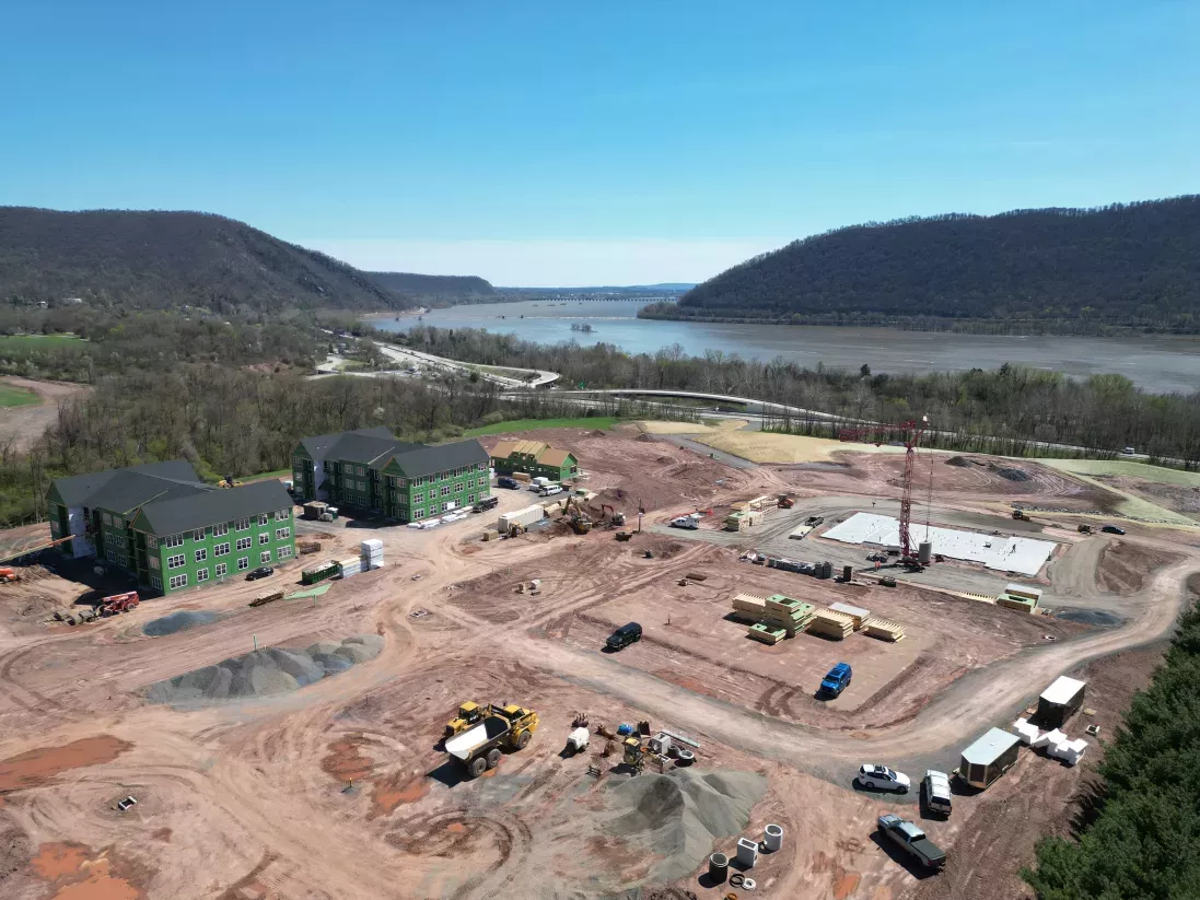 Aerial view of a large construction site with multiple buildings under construction, equipment,  a wide river, and green hills.