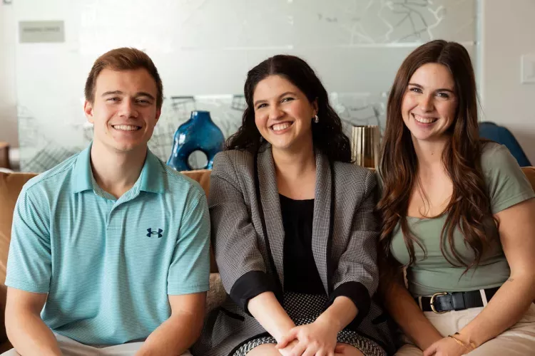 Three smiling young adults, a man and two women, sit side-by-side. A map graphic is in the background.