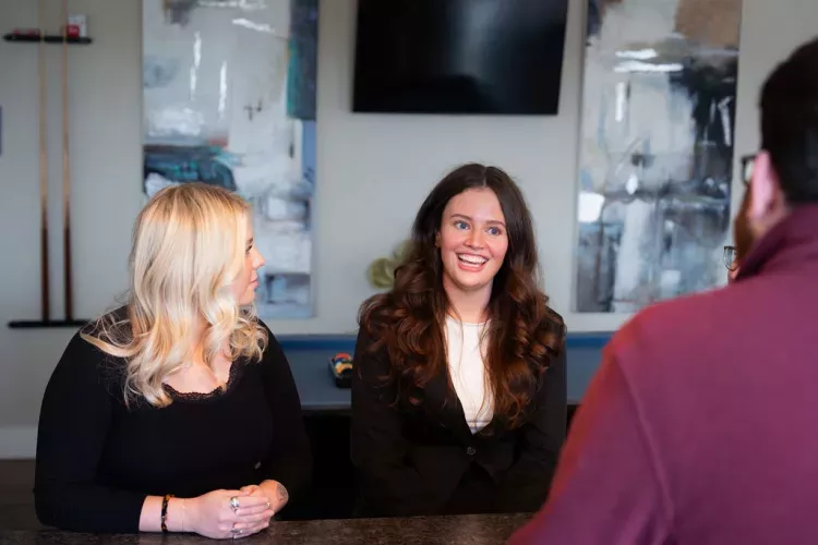 Two smiling women chat with a man across a counter in a modern lounge with abstract paintings in the background.