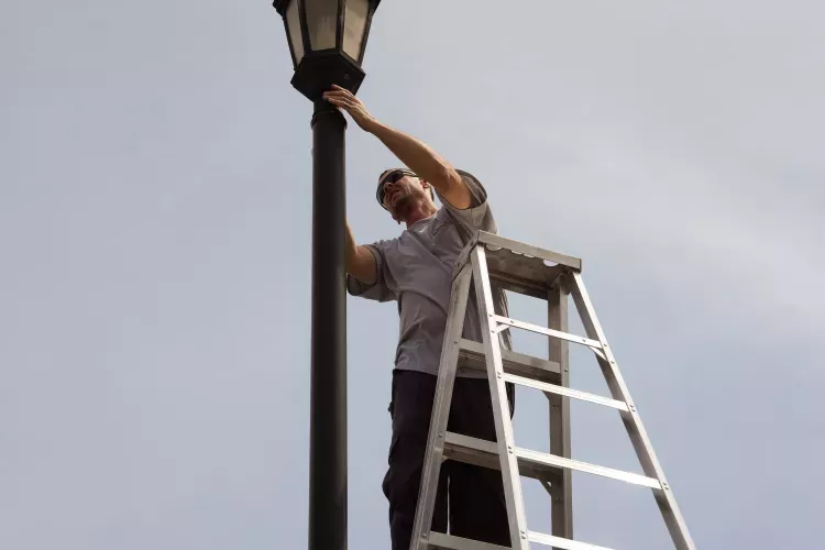 Man on a ladder adjusts a black lamppost against a clear sky.