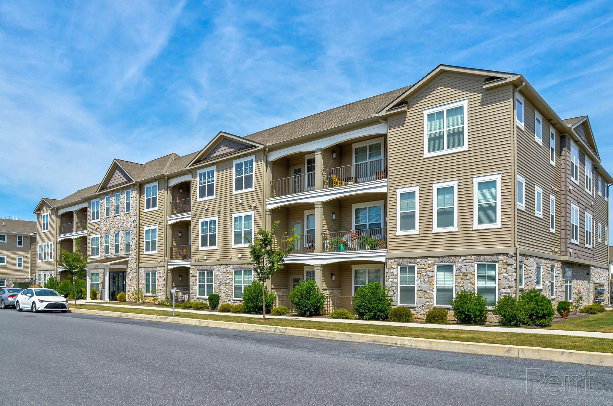 Exterior view of The Village of Old Hickory Apartments, showcasing a three-story tan and stone facade with white trim. Spacious balconies line each level, and large windows allow for natural light. Manicured shrubs and young trees enhance the curb appeal, while a quiet street with parked cars frames the front of the building.