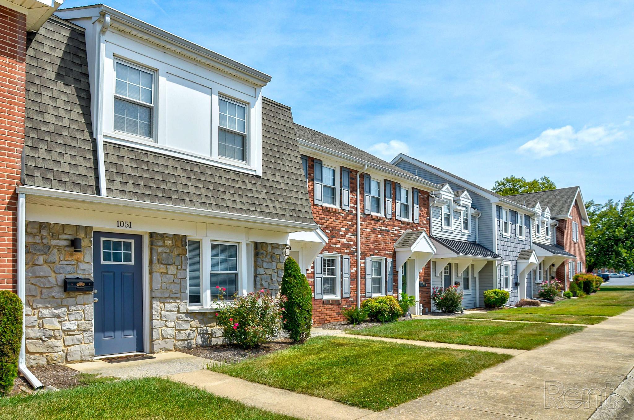 Row of brick and siding townhomes with gabled roofs and private entrances at Village of Olde Hickory Apartments, set along a clean sidewalk with manicured lawns and blooming shrubs.